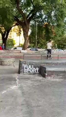 A man in a black t-shirt and dark pants skateboards in a concrete skatepark area. The man, who wears a hat, is shown jumping onto a concrete block with white “FULL SPORT” graffiti on its side, then grinding along the top of the block before successfully jumping off at the end. Other people, some with skateboards, can be seen in the background. Behind the skatepark are multiple tall buildings, some with graffiti on their lower portions. There are also trees, benches, and bikes in the background.