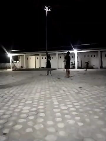 Two individuals are visible at night in an outdoor, paved area. One person, wearing a dark outfit, is standing and holding a tall, thin pole vertically with both hands. The pole extends upwards, and a bright, star-shaped object is affixed to its top, emitting light. Another person, also in dark clothing, stands to the right, observing. The background features a low, white building with multiple doors or openings and interior lighting. The ground is covered in light-colored hexagonal paving stones. The scene is illuminated by artificial lights from the building and the object on the pole.