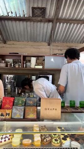 In a cafe setting, likely in Ban Sai Kham, Thailand, two individuals are engaged in preparing drinks behind a counter. The cafe features a rustic, open-air design with a corrugated metal roof and exposed walls.

The main subjects are two people wearing light-colored shirts. One, with dark, curly hair, stands facing away from the camera, seemingly operating equipment. The other, with their back to the camera and hair tied up, is leaning over the counter, engrossed in a task.

On the counter in the foreground, there is a display of various pastries and cakes. Adjacent to this display are several colorful boxes, possibly containing coffee or tea, with text that appears to be in Thai. One of these boxes is prominently labeled "KAFF CHOKTID". A wooden sign with the word "GRAPH" is also visible on the counter.

Behind the counter, shelves are stocked with various items, including what appears to be kitchen equipment and supplies. A fan is mounted on the corrugated roof. The overall atmosphere suggests a busy but relaxed environment, possibly during daylight hours given the illumination. The scene captures the everyday operations of a cafe, focusing on the preparation of beverages and the display of its culinary offerings.