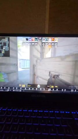 The video shows someone playing a first-person shooter game, likely Counter-Strike, on a Gigabyte laptop with a blue-lit keyboard. The game is displayed on the laptop screen, showing a scene with a light, urban background. The player is using a rifle, aiming and firing at opposing players visible in the virtual environment. On-screen elements display in-game information such as the score, time remaining, weapon stats, and the player's health.