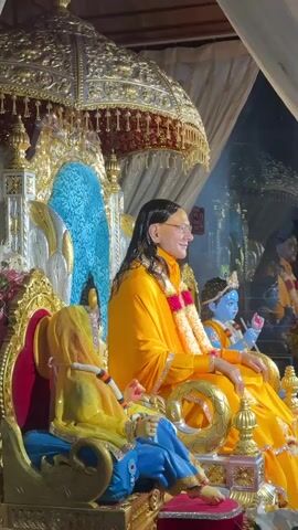 A person in a bright yellow robe sits on an ornate golden throne next to a small blue figure. To their left, another figure is adorned in yellow. The throne is elaborately decorated with gold accents, and above it sits a canopy of gold and red. Behind the figures is a reflective surface with a glimpse of people and flowers. The overall atmosphere suggests a religious or ceremonial setting. The person in yellow is wearing a garland. The throne has a blue back.