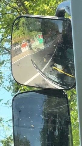 The video shows a dual-mirror setup on a vehicle, likely a bus or truck, with a view of the road behind. The upper mirror reflects a two-lane road with a white line dividing the lanes. The view in the mirror changes as the vehicle moves, revealing roadside advertisements with red and white colors, as well as green foliage. The lower mirror is more obstructed and shows scratches and dirt on its surface, limiting visibility. The sky is visible through the vehicle's windshield, and some branches of trees are seen in the foreground, adding to the sense of movement. The video is shot from inside the vehicle, offering a driver's perspective.