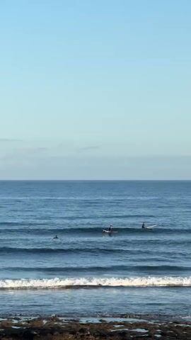 The video shows a calm ocean with surfers in the water. The sky is a clear light blue with a few faint clouds on the horizon. The waves are small and rolling towards the shore, where the rocky coastline is visible in the foreground. There are at least four surfers in the water, scattered across the frame. One surfer appears to be paddling, while others are sitting on their boards. The overall atmosphere is peaceful and serene.