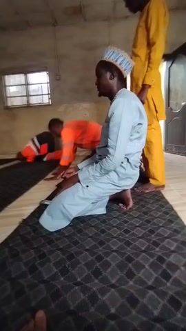 Here's a detailed description of the video:

**Scene:** The video shows a room with tiled floors and plain, painted walls. A window is visible on the left side of the frame. There's a door in the background on the right.

**People:** There are three men visible in the video.
    *   The man in the center is kneeling on a patterned rug, dressed in a light blue outfit and a traditional blue and white cap.
    *   A man standing in the background, to the right, is wearing a yellow outfit.
    *   The third man in the background, kneeling in the front left corner, is wearing a black and orange safety suit.
    *   Someone, possibly the videographer is wearing blue.

**Actions:**
    *   The three men are in the act of praying.
    *   The man in the blue outfit is kneeling in prayer, with his hands resting on his knees.
    *   The man in the orange vest is bowing towards the floor in prayer.
    *   The man in yellow is standing and appears to be in the middle of prayers.

**Objects:**
    *   A patterned rug is spread out on the floor.
    *   A window provides light to the room.
    *   A closed door is visible in the background.

**Environment:** The room appears to be a simple, functional space, possibly a room used for prayer.