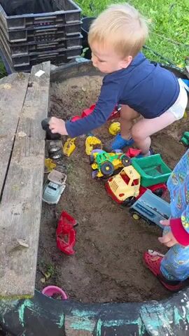 A young child with blond hair and wearing a dark blue long-sleeved shirt and a diaper is playing in a sandbox. The child is kneeling and interacting with various toy vehicles, including a black car and a red truck. Other toy vehicles and sand tools are scattered around the sandbox. The sandbox has wooden borders and appears to be outdoors, with green grass and foliage visible in the background. The child picks up a blue toy scoop. Another child's legs and feet wearing red crocs are visible in the lower right corner of the frame.