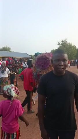 The video shows a crowded outdoor scene with a cultural event. The environment is a reddish-brown dirt surface, possibly a field or open area. Many people are gathered, including adults and children. A few individuals are wearing colorful, elaborate costumes, perhaps related to a traditional dance or performance. The atmosphere appears lively with music and movement. Buildings and trees can be seen in the background. Some individuals are walking, dancing, or watching the performers.