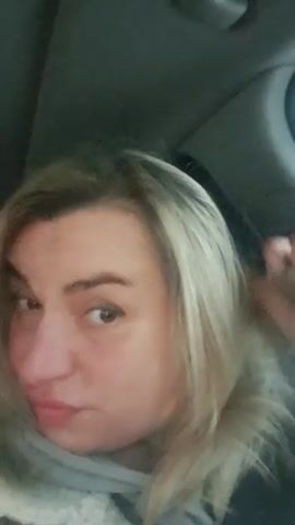 A medium shot of a woman with blond hair looking at the camera while in a car. She poses and flips her hair. The car interior is visible in the background.