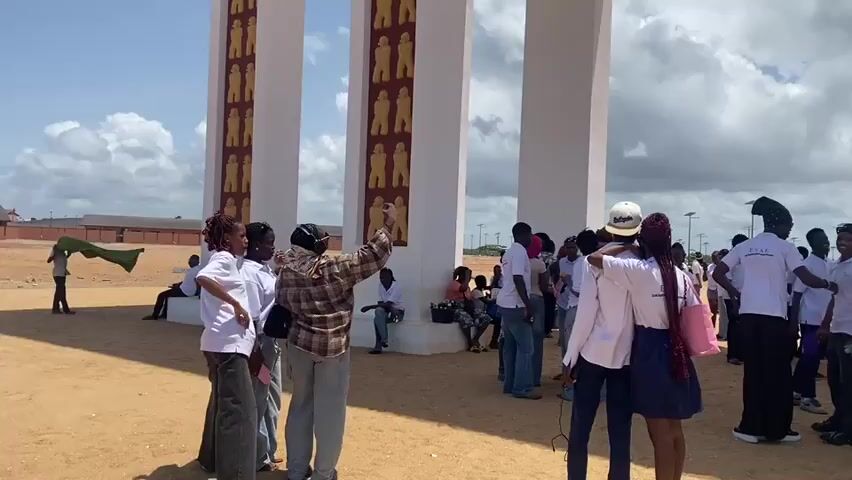 The video shows a large group of people standing in front of a monumental archway. The archway is white with reddish-brown accents and decorated with rows of golden figures on the pillars and frieze. The crowd is mostly comprised of young adults, many wearing white t-shirts with the acronym "ESAF" on them. Some individuals are taking pictures with their phones, while others are conversing or posing for photos. The ground is sandy and there's a clear view of the sky through the archway, revealing a mix of blue sky and white clouds. In the background, a long building is visible, along with several lampposts.