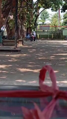 A medium shot shows a sandy outdoor area with dappled sunlight from trees. In the foreground, a blurred pink ribbon is tied around a red object. In the midground, three people are sitting on a bench near a motorcycle, and another person is riding a motorcycle in the background. A volleyball net is visible further back, behind a fence. The environment appears to be a public space, possibly a schoolyard or park.