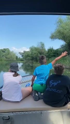 This is a video captured from the perspective of someone on a boat, looking towards the back of three individuals. The scene depicts a serene boat ride on a calm, narrow waterway bordered by lush green trees and vegetation. The sky is a clear blue with a few wispy clouds, suggesting fair weather.

The subjects are three people, two boys and a girl, all seated on the back of the boat. The girl on the left is wearing a white t-shirt, shorts, and a black bucket hat. Next to her is a boy in a bright blue t-shirt and green shorts, with his right arm outstretched as if pointing or gesturing towards something in the distance. The third person, a boy on the right, is wearing a black hoodie with a graphic of a horse and the word "Bronco" in white lettering.

The activity appears to be a leisurely boat excursion, possibly a tour or a family outing, through a natural water environment. The water is calm and reflects the blue sky and surrounding greenery. Water lilies or similar aquatic plants are visible floating on the surface. The boat itself has a metal railing and a padded seating area, indicating it's designed for passengers.

The time of day is likely midday or early afternoon, given the bright sunlight and clear sky. The overall mood seems relaxed and enjoyable, with the children seemingly engaged with the scenery or each other. The presence of trees and the calm water suggests a natural, possibly rural or park-like setting. The visible text on the hoodie reads "Bronco". The location context provided is Prevlaka, Montenegro.