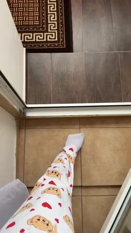 The video begins with a close-up shot of a person's legs wearing white socks and pajamas with a pattern of cartoon bears and hearts. The person is standing on a tiled floor, with a rug visible in the upper left corner. The camera pans up slightly, showing a doorway and a wall. The video then transitions to a wider, out-of-focus shot of a playground visible through a window, with apartment buildings in the background. The playground appears to be wet, possibly from rain.