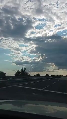 The video shows a first-person perspective of driving on a highway. The camera is positioned to capture the road ahead and the sky. The highway is paved with asphalt and has white lane markings. Guardrails are visible along the side of the road, and trees and vegetation line the edges. The sky is filled with dramatic clouds, with patches of blue sky peeking through and sunlight breaking through in some areas, suggesting either sunrise or sunset. The overall impression is of a scenic drive on a potentially overcast day.
