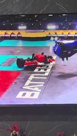 The video shows a robot combat event. Two robots are in an arena. One robot, primarily red and black, is lying on its side. The other robot, primarily blue, is in mid-air, appearing to have been thrown. The blue robot is rotating and has some damage visible. The arena floor has markings, including the word "BATTLE" visible. The background shows spectators or a stadium setting with yellow barriers and bright lights. The video is from a screen recording of the event, as evidenced by the screen's border and a partial view of figures below the screen.