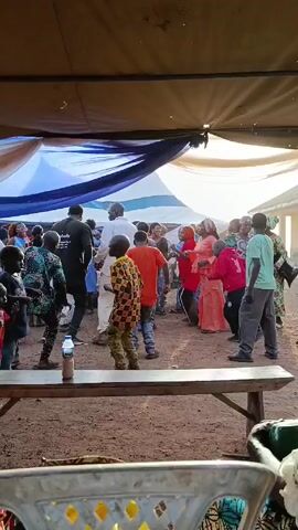 The video shows a group of people gathered under a makeshift tent, possibly for a celebration or event. The ground is dusty and covered with sand. There is a long wooden bench in the foreground with a few people sitting or standing around it. Several children are dancing and moving around in the center of the space, while others are watching or filming them with their phones. The tent is made of fabric in shades of blue, white, and brown, providing shade from the sunlight. Some adults are dressed in traditional clothing. The overall atmosphere seems festive and lively.