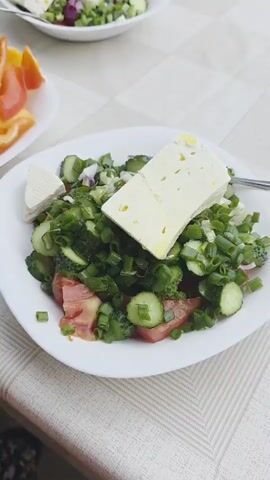 The video pans across a table with three white plates of food. One plate contains yellow and red bell pepper slices. The other two plates contain green salads with blocks of white cheese on top. The table is covered with a white tablecloth and is set in a garden. In the background, there is a green lawn, some chairs, and a watering can. No people are visible in the video.
