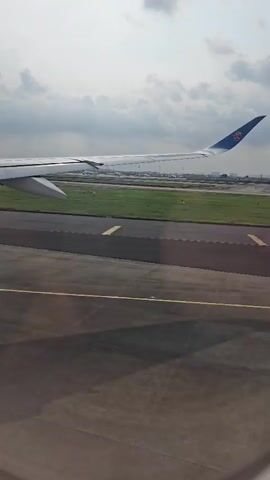 The video presents a view from inside an airplane, looking out of a window. The main subject is the white wing of the aircraft, which occupies a significant portion of the left side of the frame. In the background, the blue tail fin (vertical stabilizer) of the airplane is visible, adorned with the distinctive red and blue "Kapok flower" logo of China Southern Airlines.

The aircraft is shown taxiing on a dark grey asphalt or concrete runway, marked with prominent yellow lines. To the right of the runway, a wide expanse of green grass is visible, separating the current taxiway from another parallel runway or taxiway in the distance. Further in the background, faint outlines of airport buildings and other structures can be discerned. The sky above is overcast, filled with light grey clouds, indicating a cloudy but bright daytime. No people are visible in the frame. The scene captures the airplane in motion on the ground at an airport, consistent with either preparing for takeoff or having recently landed.