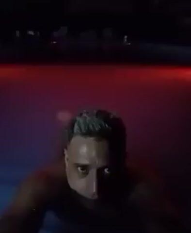 A shirtless man with short, graying hair is in a pool at night. Initially, the water is illuminated with a red light. He looks directly at the camera with a serious expression. The water then transitions to a green light. The environment is dark with faint structures visible in the background. The camera remains steady on the man's face as he remains in the water.