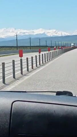 The video shows the view from inside a car driving on a highway. There are several cars ahead of the car taking the video. There is a median barrier with red markers between the lanes of traffic. Snow-capped mountains are visible in the distance against a clear blue sky. There are power lines along the side of the road.