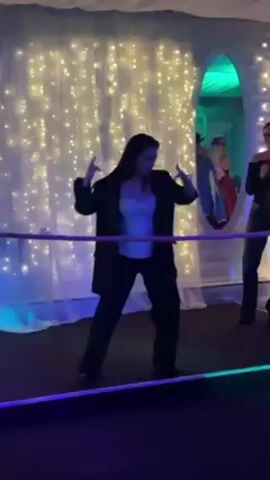 A woman in a black suit and white top is dancing in a room decorated with fairy lights and a mirrored tunnel.  She moves with energy and enthusiasm, taking off her jacket at the start and squatting low to the ground.  Several people stand nearby, observing and clapping.  The lighting is predominantly blue. A rope, lit with blue light, surrounds the dancing area. The room looks like a party or event space.