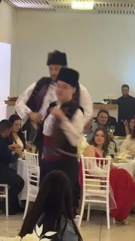 Inside a brightly lit event space, dancers wearing traditional costumes perform as seated guests observe. Two men in black hats, vests, and white shirts, one woman in a green headscarf, vest, embroidered shirt, and full skirt, and another woman in a green headscarf, vest, and white shirt dance. Guests at tables include women in dresses and men in dark shirts or suits. Some guests hold glasses and silverware. Tables are decorated with white flowers. The space is enclosed with white walls. The camera stays level, focusing on the dancers.