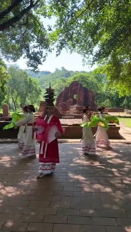 A group of five women in traditional Vietnamese attire, featuring red and white dresses and ornate headdresses, are performing a fan dance in front of ancient brick ruins. The dancers hold large pink and green fans, which they open and close in unison as they move. The scene is set outdoors in a lush green environment, with sunlight filtering through the trees overhead, casting dappled shadows on the brick pathway where the dancers are positioned. The ruins, partially overgrown with greenery, appear to be made of red brick and have intricate architectural details, suggesting a historical site. The overall atmosphere is serene and cultural, highlighting a traditional performance in a significant historical setting.