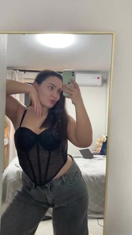 A young woman is posing for a mirror selfie in a room. She is wearing a black, see-through corset top and gray jeans. She holds her phone in her right hand and uses her left hand to pose, at one point making a hand gesture of horns. In the background is a bed with a laptop and a stuffed animal on it. There is an air conditioner mounted on the wall. The mirror has a gold frame. The walls are white. The lighting is bright.