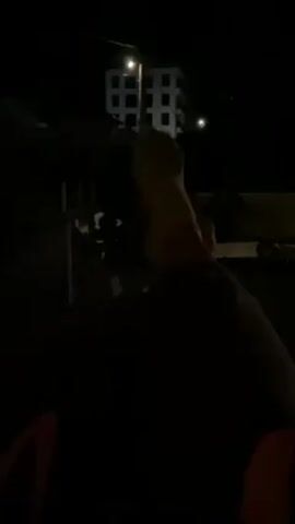 The video depicts a person at night, standing outside while smoking a cigarette. The individual's back is to the camera, with their hands visible holding the cigarette. In the background, a building with illuminated windows can be seen, along with a street light. The overall environment is dark, with minimal ambient light.