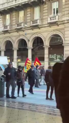 The video shows a group of people walking down a street in what appears to be an urban setting. They are holding flags, one of which is red with yellow lettering. The people are dressed in winter clothing, and some are holding phones and taking pictures. The building in the background has arched windows and appears to be quite old. There is also a white van parked on the side of the road. The overall impression is of a demonstration or protest of some kind.