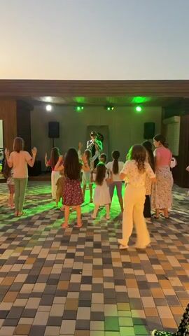 A group of children are dancing on a patterned tile floor in front of a stage where a person in a cowboy hat is singing. The stage is lit with colored lights, and there are speakers on either side. The background appears to be an outdoor venue with a wooden roof structure. The children are facing away from the camera, engaged in the performance. The lighting suggests it is either dusk or evening.