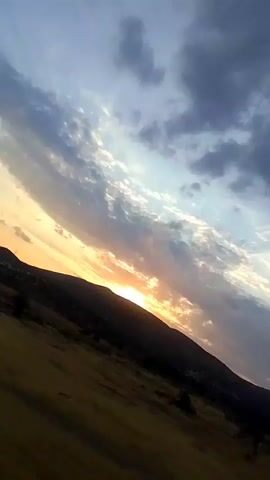 The video shows a sky with clouds and a sunset. The camera is tilted, and the sky takes up most of the frame. In the lower part of the frame, there is a dark silhouette of a hill or mountain. The sun is visible near the horizon, casting a warm glow. The clouds are a mix of white and gray, with some illuminated by the setting sun. There is also a hint of yellow and orange in the sky, indicating the colors of the sunset. In the lower right corner, there is a glimpse of trees and some man-made structures, possibly buildings. The overall scene is one of a natural landscape at dusk.
