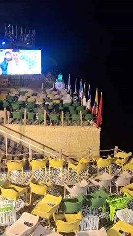 The video shows an outdoor seating area at night, possibly a stadium or a viewing event, with rows of empty and occupied chairs arranged on tiered platforms. A large LED screen is positioned at the top of the highest platform, displaying a vibrant image or video. In front of the screen, several flags are visible. The foreground features more chairs, some of which are brightly colored. People are seated in some of the chairs on the elevated platforms, facing the screen. The overall atmosphere appears to be that of a public gathering to watch an event.