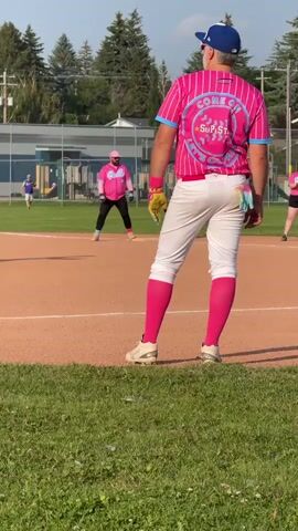 The video shows a baseball game with several players in pink uniforms. In the foreground, a person wearing a pink and blue striped baseball jersey with "COME ON SUP STAR" printed on the back, white pants, and pink socks is standing on a baseball field. The person is wearing a blue baseball cap and yellow gloves. In the middle ground, another person in a pink and blue striped baseball jersey with "BARBIE" printed on the front, a pink cap, black pants, and pink socks is standing on the field. To the left, a third person in a blue and black uniform is pitching the ball. The field has a dirt infield and grass outfield, with a fence and trees in the background. The sky is blue with some clouds. The video appears to be shot during the daytime.