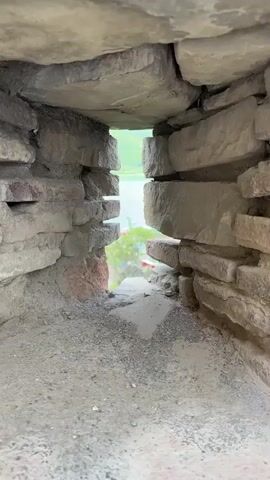 The video shows a view through a stone opening. The opening is made of rough-hewn stone blocks. Inside the opening, the ground is made of gravel and dirt. Outside the opening, there is a body of water, a green forested hillside, and a small town with red-roofed buildings. The sky is partly cloudy. The camera pans slightly, showing different angles of the opening and the landscape beyond.