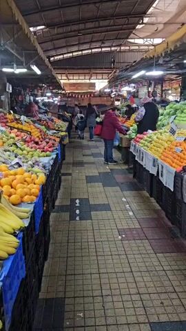 The camera slowly traverses through a vibrant indoor fruit and vegetable market. Along both sides of a central aisle with a patterned tile floor, vendors display their produce in black plastic crates and on tables covered with white paper. An abundance of fruits, including oranges, apples, pears, plums, kiwis, bananas, and pomegranates, are neatly stacked, adding to the market's colorful atmosphere. 

Several customers are observed browsing and purchasing goods. They are dressed in casual winter clothing, with some wearing jackets and carrying shopping bags. A few vendors are visible behind their stands, interacting with customers. The market is covered by a high, arched metal roof, supported by visible beams and illuminated by fluorescent lights. Banners and decorations add a touch of festivity to the market's ambiance.