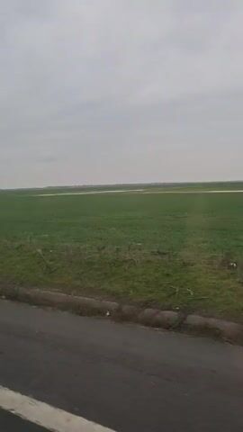 The video shows a view from the window of a moving vehicle, likely a car. The focus is on the exterior scenery, which is primarily a flat, green field under a gray, overcast sky. The road is visible in the foreground, with a white line marking the lane. As the car moves forward, a roadside billboard with the word "GECOR" is visible on the right side of the road. A bare tree is also seen near the billboard. The video appears to be taken during the day, with no distinct people or interactions visible. The audio contains music.