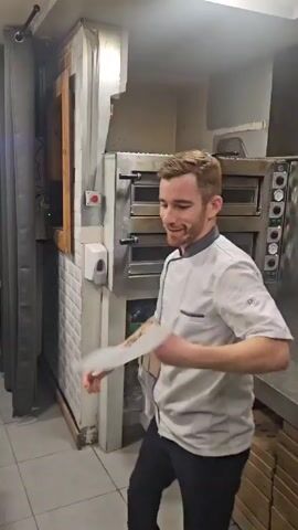 A chef is seen in a brightly lit kitchen. He is wearing a short-sleeved white chef's coat and dark pants, with a tattoo visible on his left arm. The chef is tossing and spinning a round, flat piece of white dough into the air, seemingly practicing pizza-making skills. He is smiling and looking towards the camera. Behind the chef are industrial kitchen appliances, including multiple ovens. The kitchen has tiled floors and walls with various equipment and supplies stored around.