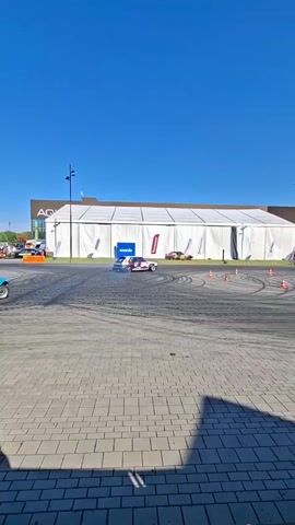 Two drift cars perform donuts on a paved area in front of a large white tent structure. The cars emit plumes of smoke as they slide sideways, leaving tire marks on the pavement. Orange cones are arranged in a circular pattern on the ground. The sky is clear and blue.