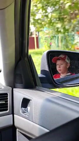 A young boy wearing a red baseball cap and a red shirt is visible in the passenger side mirror of a vehicle. He appears to be looking around, possibly at the passing scenery. The background outside the car shows a rural setting with houses, trees, and a dirt road. The car appears to be in motion.
