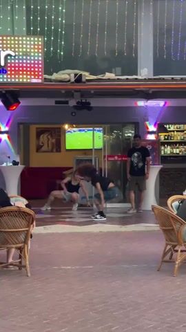 At approximately 00:00:00, a man performs a backflip in front of a bar. A woman in the foreground is doing a plank exercise and a second woman is in a lunge. Two men stand watching the performance. The bar has colorful lights and a television screen displaying a soccer game. People are seated at tables in the foreground, some of whom are looking at the performance. At 00:00:03, a man walks past the scene. The lighting suggests it is nighttime.