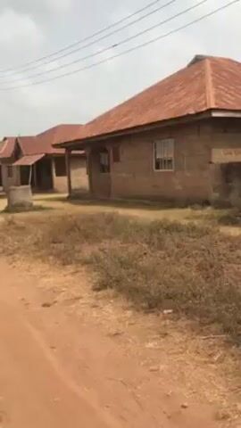 The video shows a row of simple houses along a dirt road, likely in a rural area. The houses are made of block construction with rusty, corrugated metal roofs. Each house has a small covered porch area. The grounds are dry and covered with sparse, brown vegetation. Overhead power lines are visible. The sky is overcast. There is no evidence of human activity in the video.
