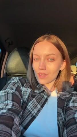 Here's a description of the video:
The video shows a fair-skinned woman with shoulder-length light brown hair in the driver's seat of a car. She wears a brown and grey plaid shirt over a white top, and gold chain earrings. She is wearing a silver ring on her left hand and a small cross pendant on a gold chain around her neck. She looks directly at the camera and smiles. Sunlight streams into the car, illuminating her face. In the background, visible over the passenger seat, is the blurred image of another person. The background shows sky and a field through the window. The camera is held at a selfie angle.