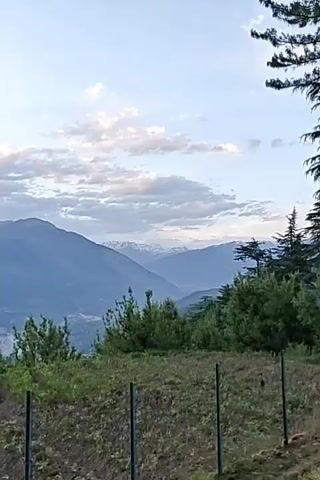 The video shows a landscape with mountains in the background. Some snow-covered peaks are visible in the distance. The sky is partly cloudy with a mix of blue and gray. In the foreground, there's a field with some green vegetation and a partially visible fence. Some trees are scattered around the edges of the field.