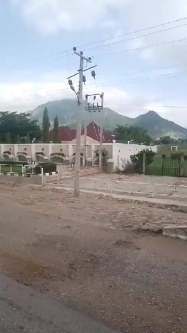 This media file captures a scenic view of a long, white building with arched windows, likely a residential or commercial property, situated in Wuro Sani, Nigeria. The building is set against a backdrop of lush green trees and rolling hills that disappear into a hazy sky. The foreground shows a dirt road, suggesting a rural or semi-urban environment. A utility pole with electrical wires stands prominently on the right side of the frame, indicating the presence of modern infrastructure. The scene is bathed in natural light, suggesting it was filmed during the daytime, possibly in the morning or late afternoon, given the soft illumination and overcast sky. The weather appears to be cloudy, with the clouds obscuring the full intensity of the sun. There are no people visible in the frame, and the overall atmosphere is peaceful and serene. The architecture of the building, with its uniform design and white facade, creates a sense of order and uniformity. The presence of manicured greenery along the fence in front of the building adds to the aesthetic appeal. The video pans slowly from right to left, offering a panoramic view of the property and its surroundings. The media file does not contain any visible text.