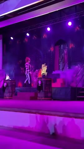 The video captures a theatrical performance on a stage bathed in purple light. A performer, dressed in a striped costume and with theatrical makeup, stands center stage on a raised platform. They are interacting with the environment, which includes smoke effects and props resembling barrels with mask-like decorations. To the left and right of the stage, there are also elements that appear to be part of the set design, possibly representing a dark or spooky theme. Another figure is visible in the background on the left, and a tall, silhouetted figure stands on the far right. The overall atmosphere is dramatic and visually striking, with the colored lighting playing a significant role in setting the mood.