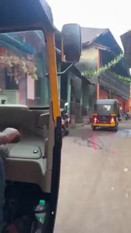 The video is taken from inside a yellow and black auto rickshaw, moving on a narrow, brick-paved street. The driver's hand is visible on the steering column. A water bottle sits on the floor. Other vehicles, including another rickshaw and a motorbike, are visible moving in the same direction. Buildings line both sides of the street, varying in color and architectural style, with some appearing old and possibly residential. Some people are walking on the side of the street. The weather appears overcast, and the road surface is wet.