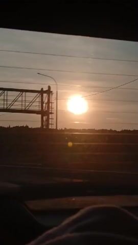 The video shows a view from inside a vehicle, looking out the rear window at a sunset. The sun is bright and appears to be low on the horizon. There are power lines visible across the sky, and a silhouetted landscape with some structures is in the distance. The bottom of the frame shows a blurry portion of a person's leg or lap, indicating the perspective of someone seated in the vehicle. The overall lighting suggests it is either sunrise or sunset.