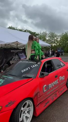 Two women wearing green and black outfits are sitting on top of a red car. The car has "Ceramic Pro" and "Drift Taxi" written on it. There is a white tent and a group of people in the background. The sky is cloudy.