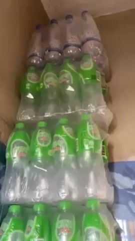 A stack of unopened, plastic-wrapped packs of green soft drink bottles is visible. At the very top, a stack of clear, plastic-wrapped bottles is partially visible.  The bottles are stacked against a wall that is cream colored and adjacent to a wall with blue and white tiles.