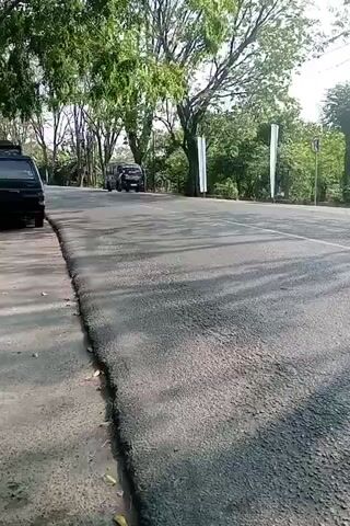 The video shows a road with several motorcycles and one car. The road is paved and has a sidewalk on the left side. There are trees lining the road, and the sky is visible above. In the foreground, the pavement is rough and cracked, with some debris on the side. A dark colored car is parked on the left side of the road. Several motorcycles are driving on the road, some with riders wearing helmets. One motorcycle with a rider wearing a red helmet is clearly visible in the center of the frame, moving away from the camera. The lighting suggests it is daytime, possibly late afternoon due to the long shadows cast by the trees.