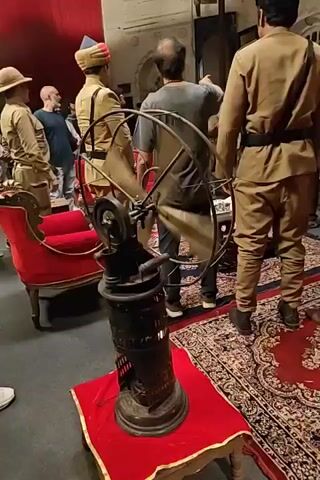 The video shows a vintage fan operating on a small table, adorned with a red cloth. The fan is dark in color, with a wheel-like structure at the top and a cylindrical base that has a visible flame burning inside. Several people are in the background, some wearing what appears to be uniform-like clothing, one in a hat. The room seems to be staged with older furniture, including a red-upholstered chair, and a patterned red rug on the floor. A few other people can be seen standing around, observing the scene.